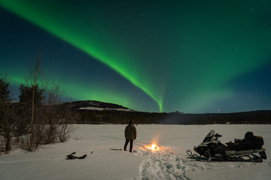 Het noorderlicht bij Friluft Sweden. Een sneeuwscooter en vuurtje op het ijs net groene noorderlicht strepen in de lucht.